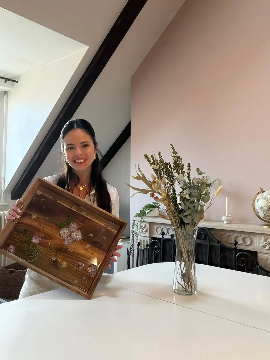 Amanda Berton souriante, tenant un plateau garni de fleurs pressées dans un intérieur clair
